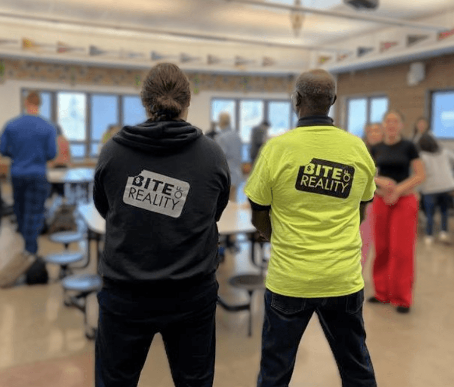 Two adults standing indoors wearing Bite of Reality shirts with people and tables in the background